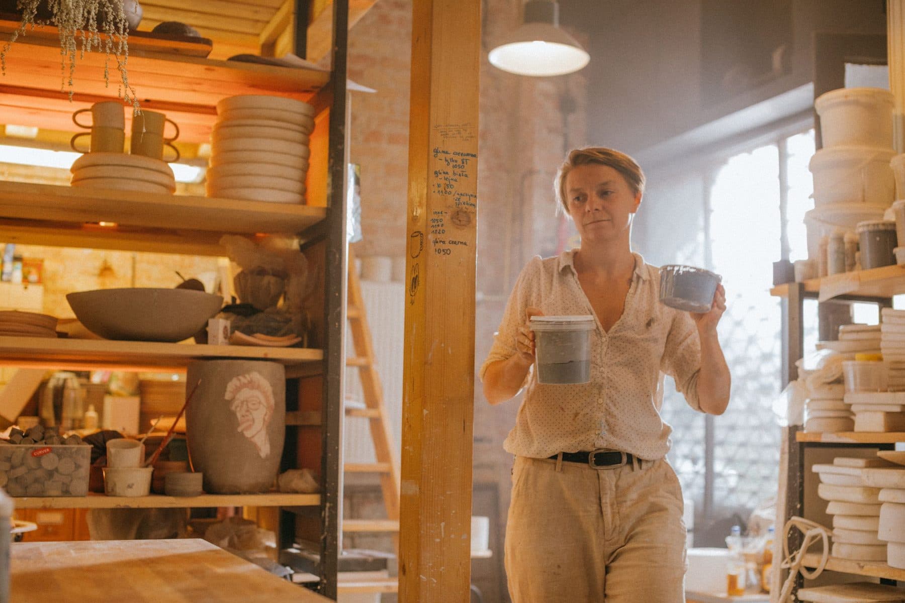 Une céramiste porte des bols dans son atelier, entre les étagères de poteries.