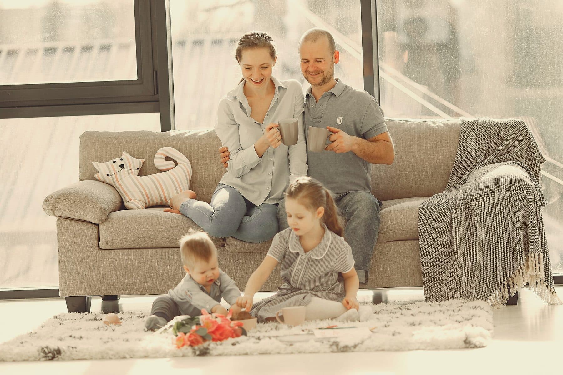 Des parents sur le canapé de leur salon, leurs deux jeunes enfants jouent sur le tapis.