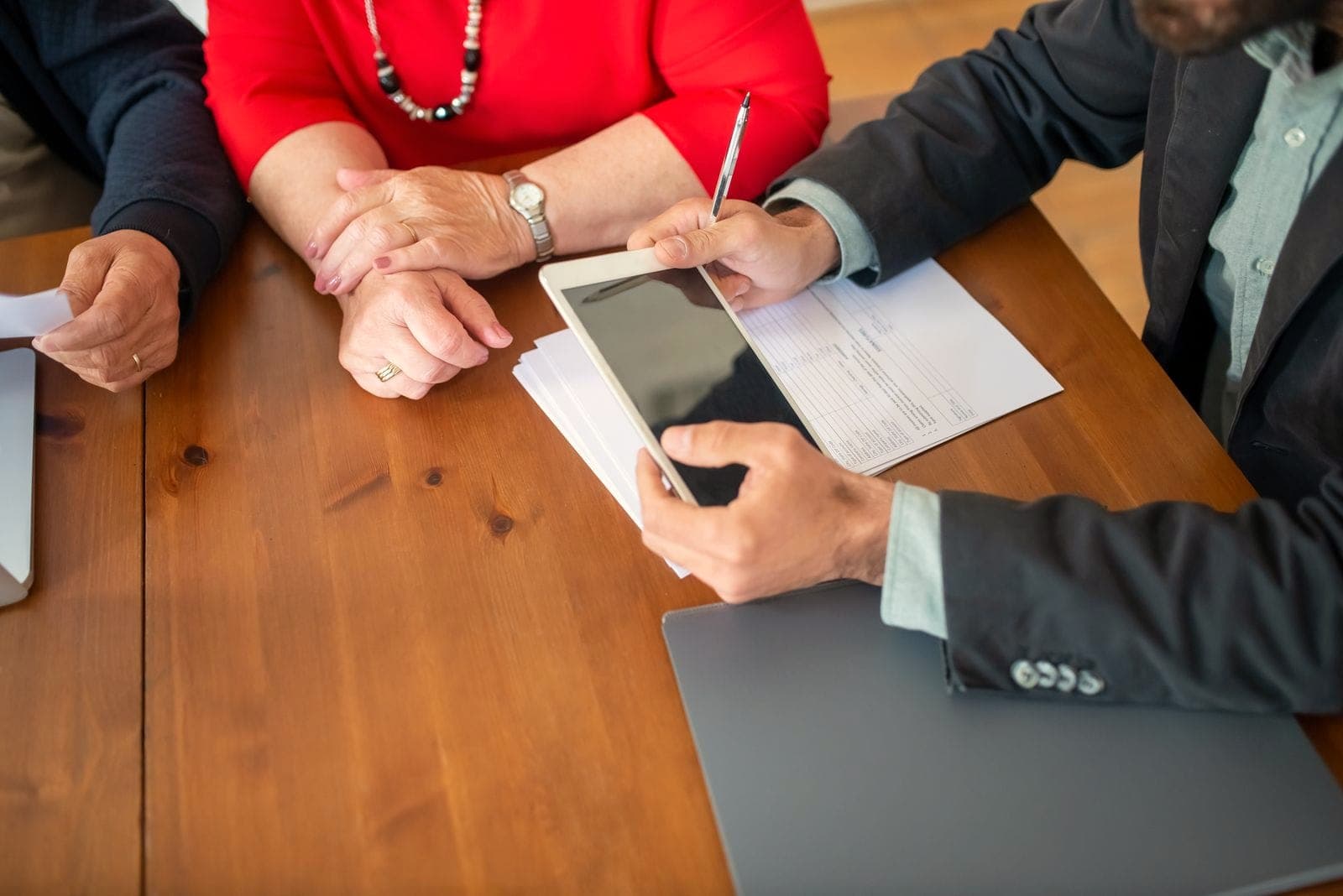Des mains autour de documents et d'une tablette, de part et d'autre d'une table en bois.