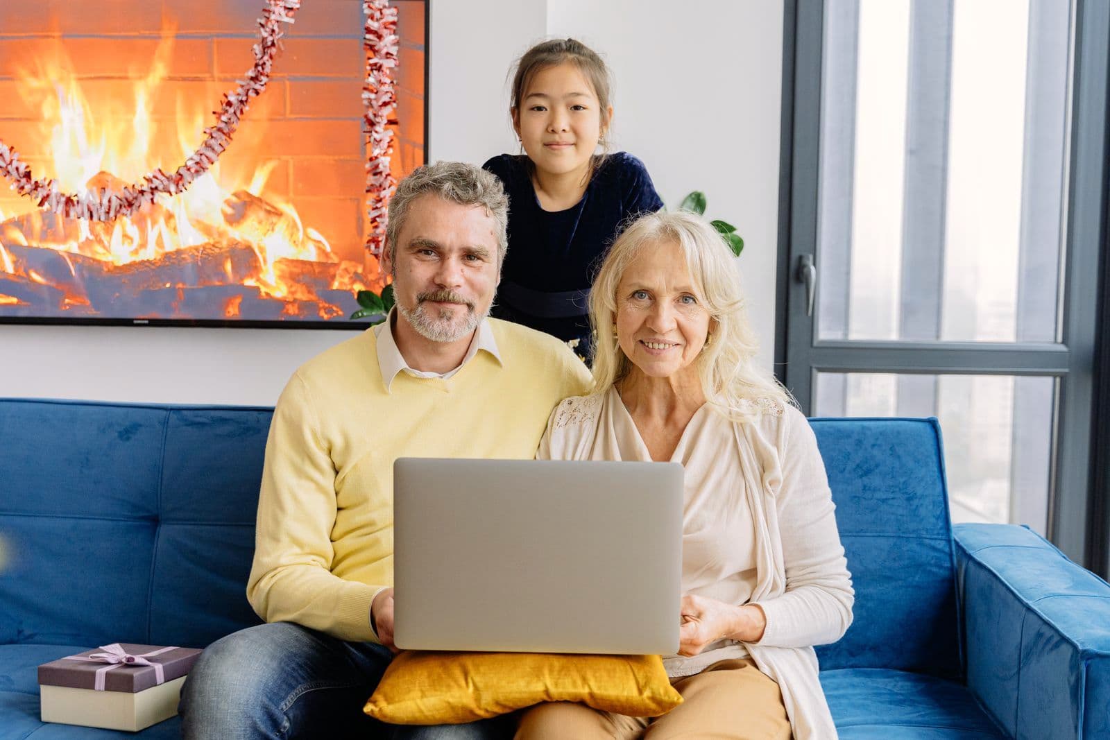 Des grands-parents et leur petite-fille regardent ensemble un ordinateur portable, sur le canapé du salon.